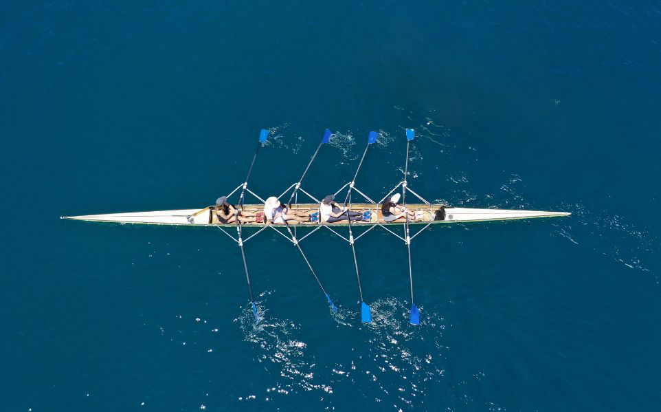 Four people rowing a racing boat in blue water, seen from above.